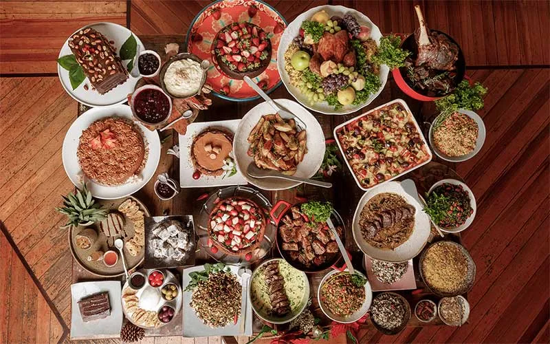 Mesa farta com pratos variados vistos de cima: assados, saladas, farofas e sobremesas