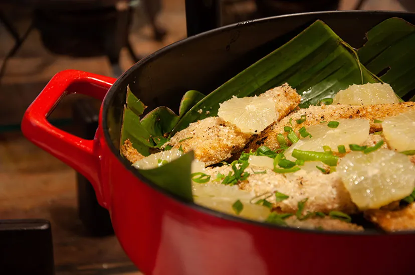 Peixe com legumes servido em panela vermelha sobre folha de bananeira